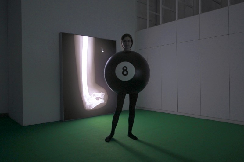 Zdjęcie: widok wystawy, przygaszone światło, jasnozielona wykładzina; pośrodku stoi kobieta w czarnych rajtuzach, tułów ukryty w ogromnej, czarnej bilardowej kuli z numerem 8; w tle lightbox, na nim rentgen kości kończyny. 