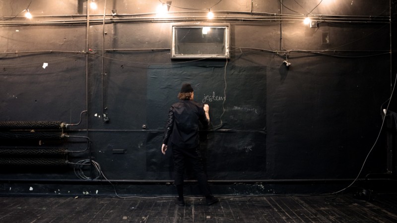 Zdjęcie: ciemne wnętrze; podłoga, ściany, oraz elementy instalacji technicznych pomalowane na czarno; na środku mężczyzna ubrany na czarno, w czapce, tyłem do nas, pisze na ścianie, powyżej napis kredą „jestem”.