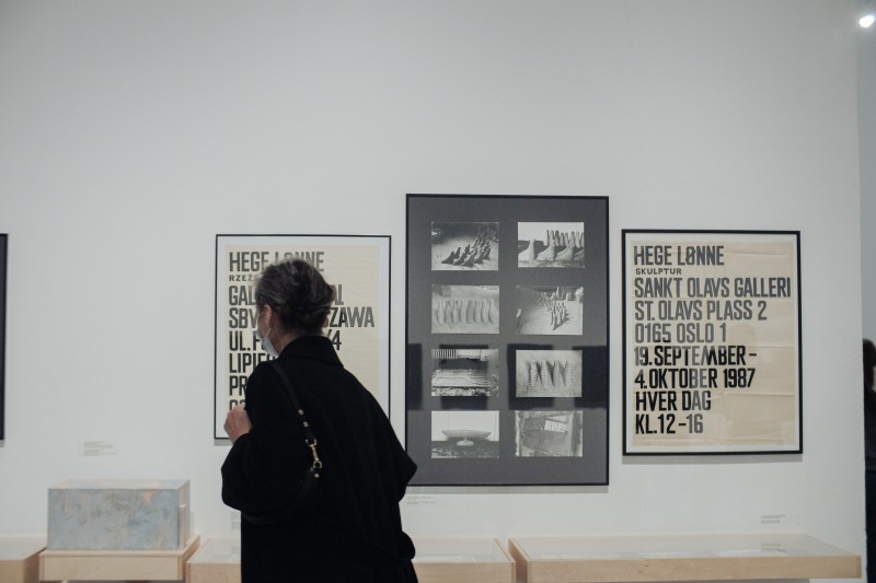 Zdjęcie przedstawiające widok wystawy. Osoba odwrócona tyłem do obiektywu patrzy na zdjęcia wiszące w dużej ramie. Obok zdjęć wiszą liternicze plakaty wystawy z 1987 roku