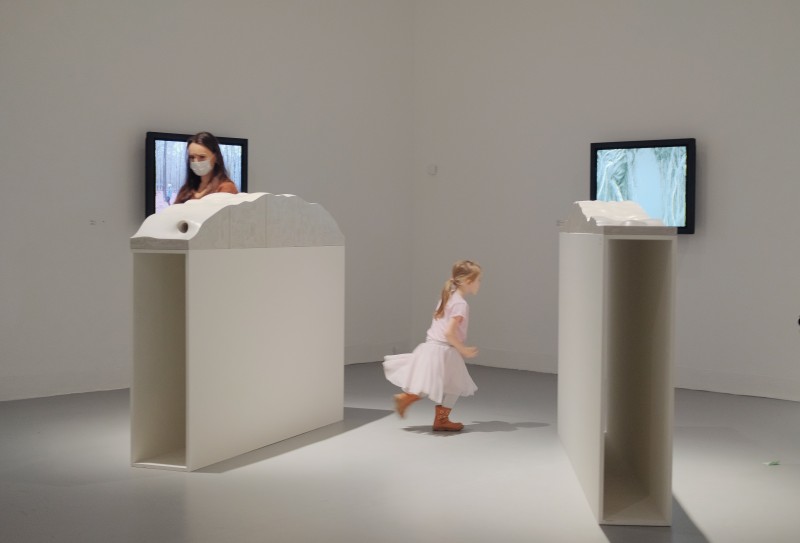 zdjęcie z wystawy, białe wnętrze, białe postumenty podtrzymujące abstrakcyjne rzeźby, na ścianie dwa ekrany. Po środku dziewczynka w  jasnoróżowej sukience biegnąca między kubikami.