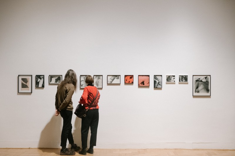 Zdjęcie: widok wystawy. Dwie osoby oglądają fotografie zawieszone na ścianie w rzędzie. Fotograwie są w różnych formatach, w czarnych ramkach. Przedstawiają fragmenty kobiecego ciała. 