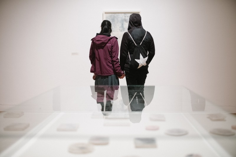 Zdjęcie: widok wystawy. Dwie osoby oglądają grafikę zawieszona na ścianie. Pierwszy plan nieostry - niewyraźnie widać niewielkie obiekty zamknięte w szklanej gablocie. Odbijają się w niej sylwetki oglądających. 
