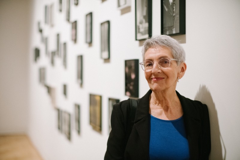zdjęcie: starsza kobieta, w krótkich, siwych włosach i okularach stoi na tle ściany, na której nieregularnie są powieszone fotografie w małym formacie