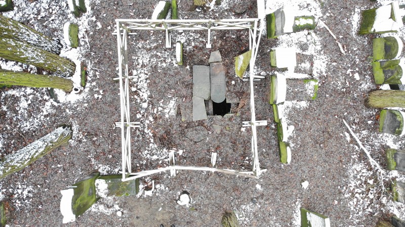 Widok z góry na nieduży fragment cmentarza, na środku otoczona płotem dziura w ziemi, częściowo przykryta kamiennymi płytami.