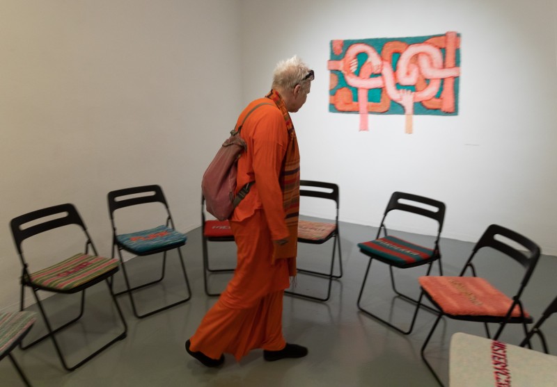 Osoba ubrana na pomarańczowo w przestrzeni galerii, za nią krzesła