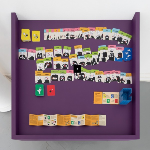 Kwadratowa, fioletowa półka; widok z góry. Na powierzchni rozłożone: kolorowe karty, instrukcja, pudełko.