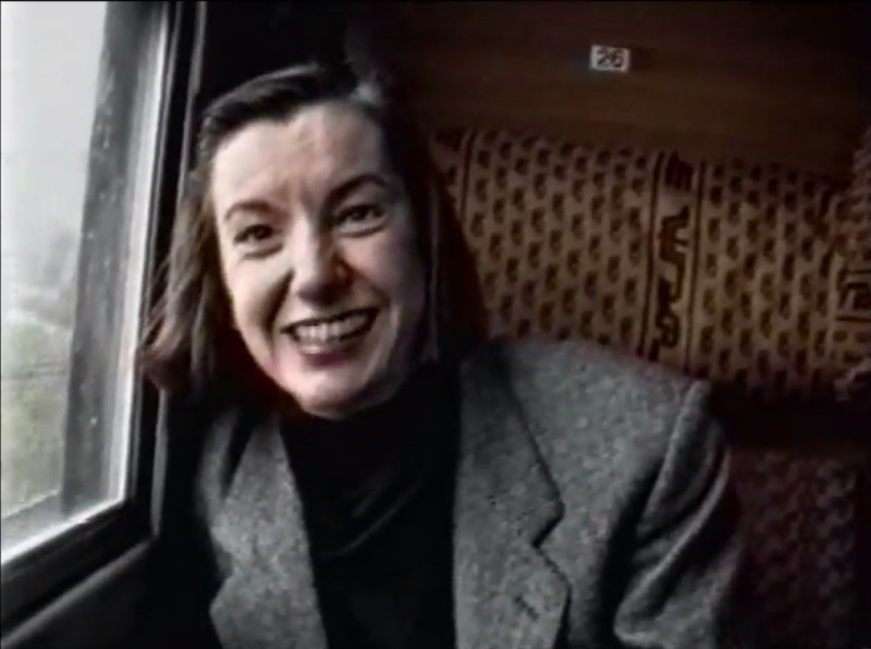 Zdjęcie o nieco przygaszonych kolorach. Wnętrze wagonu pociągu. Zbliżenie na twarz uśmiechniętej kobiety, która siedzi przy oknie.  