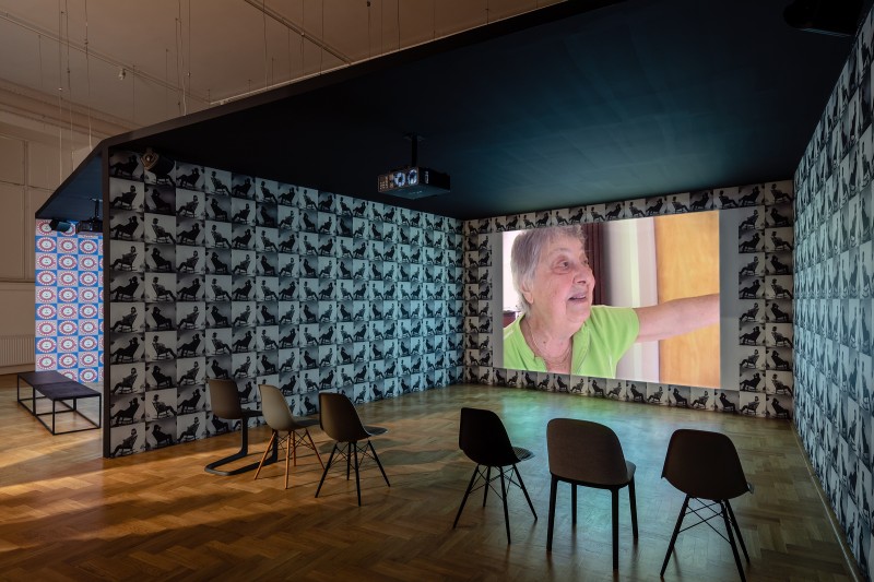 Sala z tapetą z geometrycznymi kształtami. Projekcja filmu na jednej ze ścian, na niej twarz starszej kobiety.