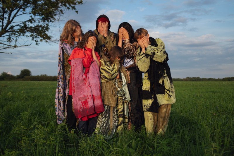 Grupa ludzi w kolorowych tkaninach stojąca na tle zielonej łąki. Osoby zasłaniają twarze dłońmi