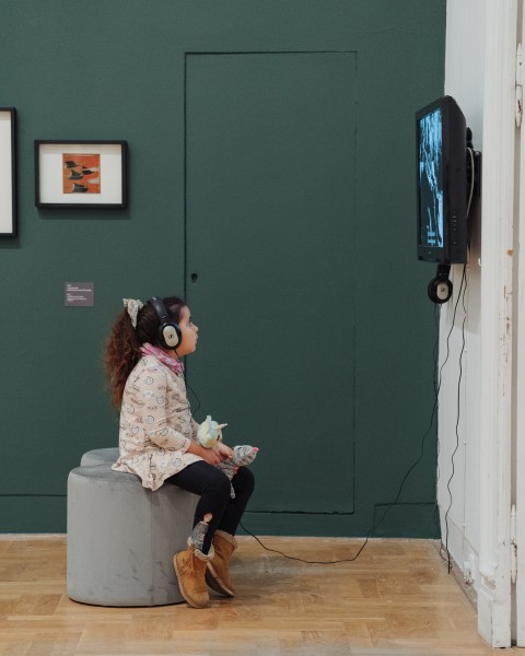 Zdjęcie. Dziecko siedzące na pufie ogląda film na ekranie w przestrzeni wystawy. W tle ciemno-zielona ściana.