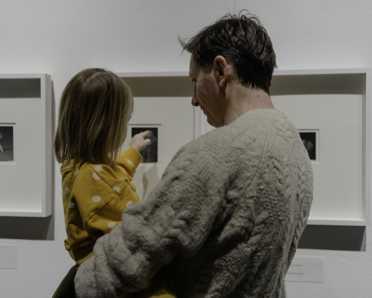 Zdjęcie. Mężczyzna trzymający dziecko na rękach przed ścianą z powieszonymi grafikami.