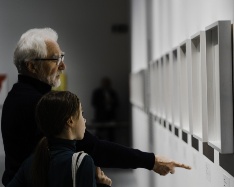 Zdjęcie. Starszy człowiek z dzieckiem przy ścianie z oprawionymi pracami. Dziecko wskazuje palcem na jeden z obrazów.