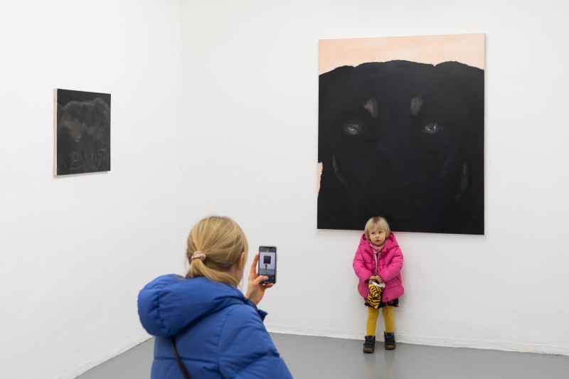 Zdjęcie. Dziewczynka w sali wystawowej na tle obrazu z psem, mama dziewczynki robi jej zdjęcie telefonem.