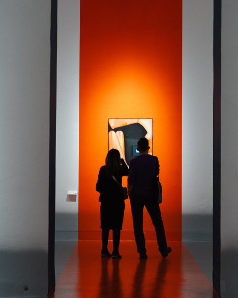 Dwie osoby w przestrzeni wystawy. Oglądają obraz wiszący na pomarańczowym tle
