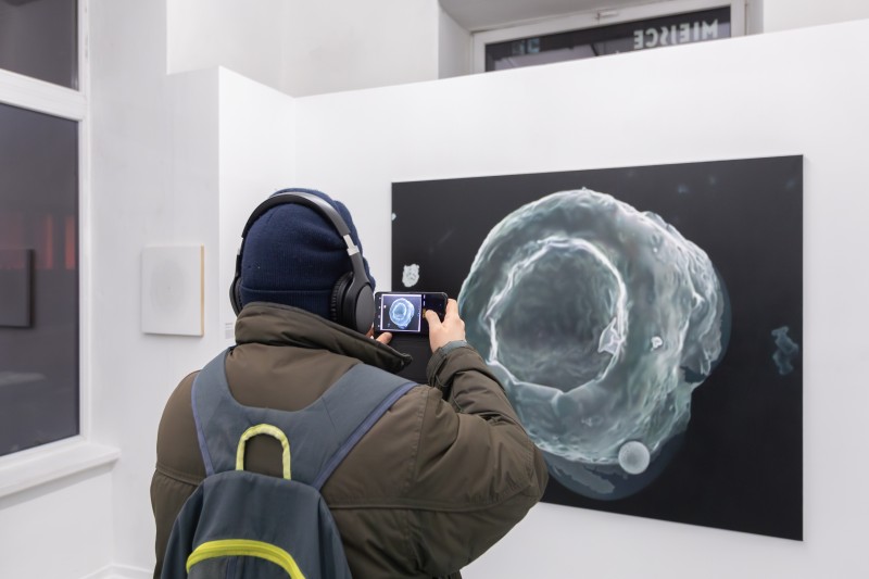 Zdjęcie, osoba w kapturze fotografuje telefonem, czarny obraz olejny na białej ścianie wystawowej. Na obrazie szara cząsteczka w powiększeniu