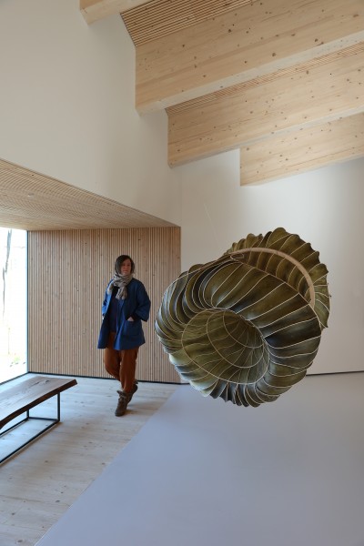 Zdjęcie; kobieta w nowoczesnym wnętrzu przechadza się koło podwieszonej cylindrycznej rzeźby o organicznym kształcie.