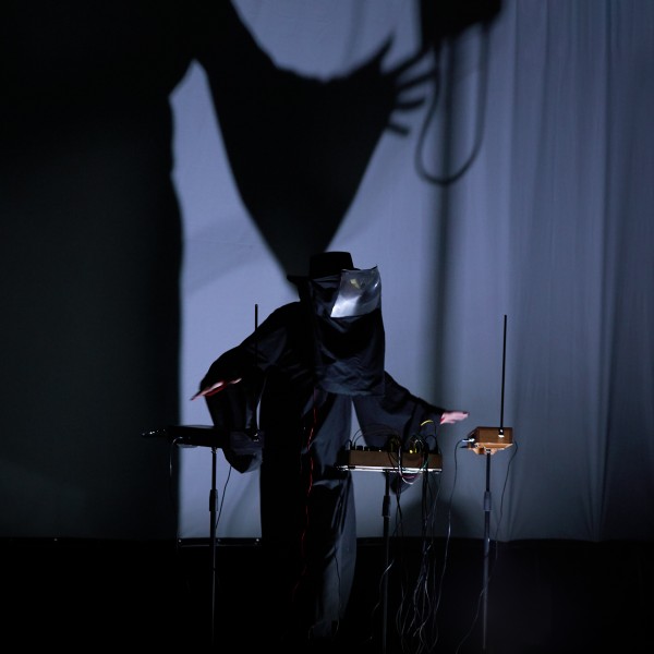 Zdjęcie, ciemne pomieszczenie, sylwetka zamaskowanej osoby nad sprzętem muzycznym.