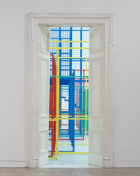kolorowe zdjęcie; kadr otwartych białych drzwi na sali wystawowej, za drzwiami monumentalna metalowa instalacja ze zespawanych elementów w kolorach żółtym, niebieskim, czerwonym i zielonym układających się w geometryczne struktury
