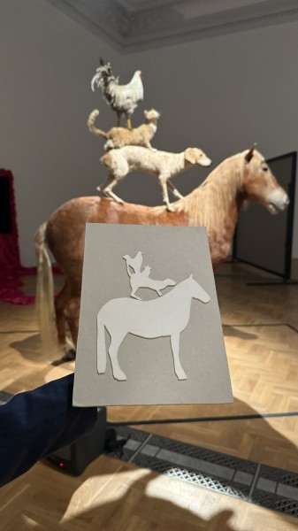  kolorowe zdjęcie; kadr na tekturową tyflografikę rzeźby PIRAMIDA ZWIERZĄT Katarzyny Kozyry, w tle oryginalne dzieło: Instalacja złożona z wypreparowanych ciał czterech zwierząt, stojących jedno na drugim: na dole brązowy koń, na nim beżowy pies, rudy kot 