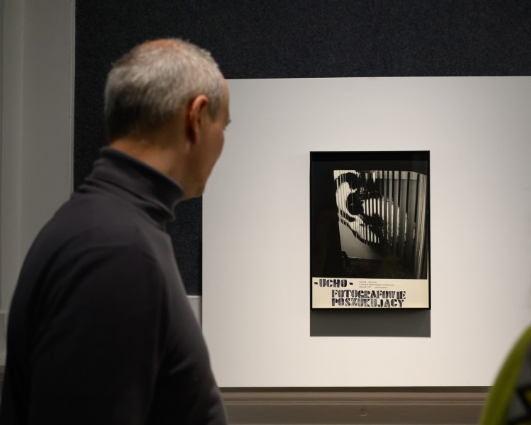 Mężczyzna stoi bokiem i patrzy na czarno-białą fotografię ucha z napisem UCHO – FOTOGRAFOWIE POSZUKUJĄCY.