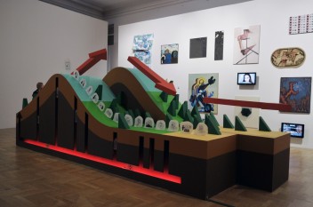 Widok sali ekspozycyjnej, po przekątnej; na środku duży, kolorowy obiekt przypominający schematyczną makietę rzeźby terenu, z wzniesieniem. Nad makietą trzy czerwone strzałki podwieszone pod sufitem sugerują kierunek od lewej do prawej. Makieta kolorowa: n