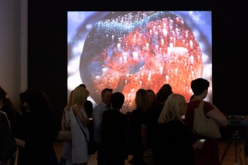 Sala wystawowa, półmrok; na pierwszym planie kilkanaście osób; w tle – wyświetlona na ekranie abstrakcyjna, kulista forma w odcieniach kolorów: niebieskiego i pomarańczowego.
