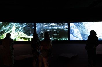 Wnętrze sali wystawowej; jest ciemno, na pierwszym planie zarys kilku sylwetek zwiedzających; w tle trzy ekrany obok siebie, na których wyświetlane są filmy: obrazy w odcieniach niebieskiego i zielonego. 