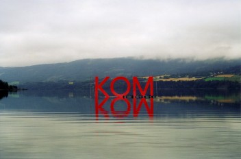Zdjęcie: Krajobraz z jeziorem na pierwszym planie oraz górzystym terenem na drugim. Wzgórza spowija mgła. Na tafli jeziora utrzymuje się czerwony napis KOM, który lustrzanie odbija się w wodzie.