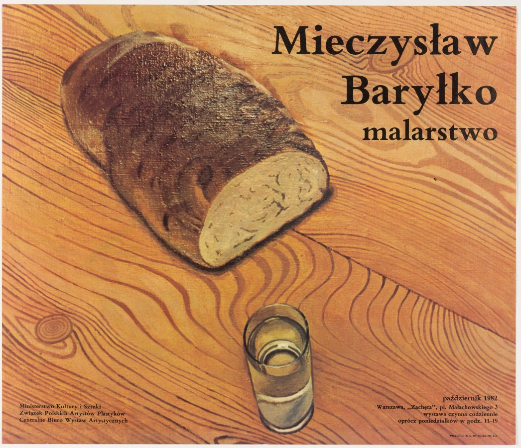 Na blacie drewnianego stołu leży chleb z odkrojonym kawałkiem, a obok stoi wypełniona do połowy przezroczystym płynem cienka szklanka. 