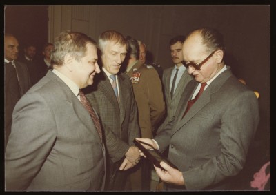 Kolorowe zdjęcie. Zbliżenie na mężczyzn w szarych garniturach. Z prawej strony generał Wojciech Jaruzelski. Przygląda się ramce, którą trzyma w rękach.