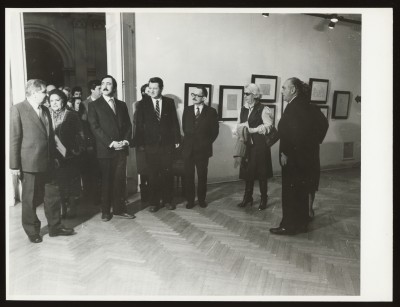 Czarno-białe zdjęcie. Kilka osób w sali wystawowej. Do sali wchodzi grupa osób z sali obok. Na ścianie wiszą oprawione grafiki.