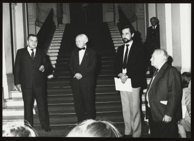 Ciemne czarno-białe zdjęcie. Czterech mężczyzn w ciemnych garniturach stoi na schodach wewnętrznych Zachęty. Jeden trzyma kartkę, zdaje się mówić.