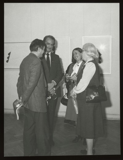 Czarno-białe zdjęcie. Dwie kobiety i dwóch mężczyzn stoją w grupie i rozmawiają, trzymają m.in kwiaty, teczkę, magazyn. W tle wiszą minimalistyczne grafiki.