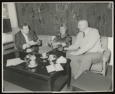Czarno-białe zdjęcie. Przy szklanym stoliku, na którym stoją filiżanki siedzą trzy osoby. Mężczyzna trzyma kartkę i długopis, kobieta i mężczyzna przeglądają gazetę