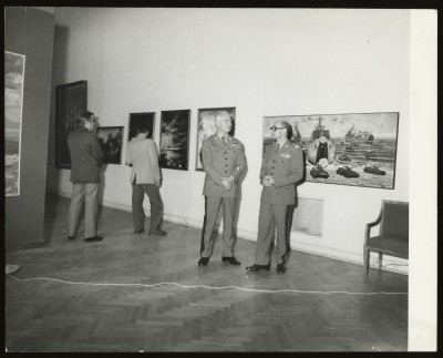 Czarno-białe zdjęcie. W przestrzeni wystawy czterech mężczyzn. Dwóch ogląda wiszące na ścianach obrazy, dwóch w mundurach rozmawia ze sobą.