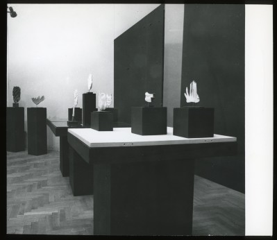 Czarno-białe zdjęcie. Widok wystawy. Na czarno-białych postumentach stoją małe postumenty, na których stoją nieduże rzeźby. W tle czarne panele.