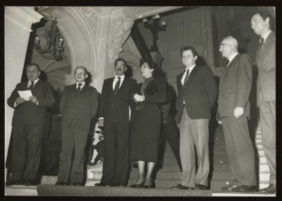 Czarno-białe zdjęcie. Sześciu mężczyzn i kobieta stojąca w środku stoją na półpiętrze wewnętrznych schodów Zachęty. W tle detale architektoniczne.