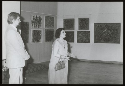 Czarno-białe zdjęcie W przestrzeni wystawy mężczyzna po lewej i kobieta w centrum. Kobieta opowiada coś gestykulując, w tle abstrakcyjne obrazy na ścianach.  