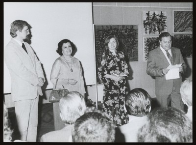 Czarno-białe zdjęcie W przestrzeni wystawy mężczyzna po lewej i kobieta w centrum. Kobieta opowiada coś gestykulując, w tle abstrakcyjne obrazy na ścianach.  