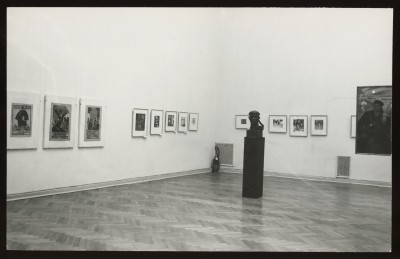 Czarno-białe zdjęcie. Widok wystawy. Na białych ścianach oprawione nieduże grafiki. Na środku sali na postumencie rzeźba: głowa Lenina.