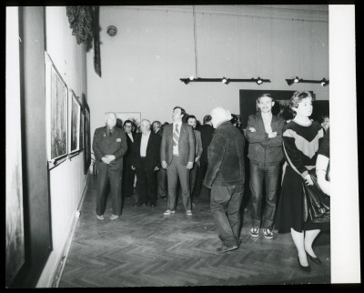 Czarno-białe zdjęcie. W przestrzeni wystawy kilka osób. Stoją w grupach i rozmawiają. W tle tkaniny wiszące na białej ścianie.