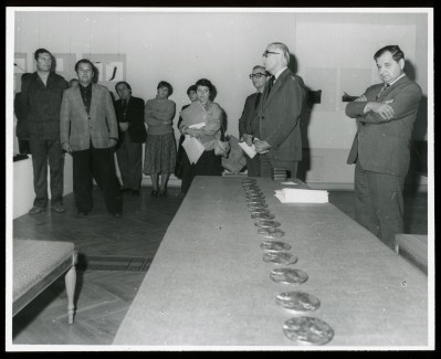 Czarno-białe zdjęcie. Kilkanaście osób stoi przy postumencie, na którym ułożone są w rzędzie spore medale.
