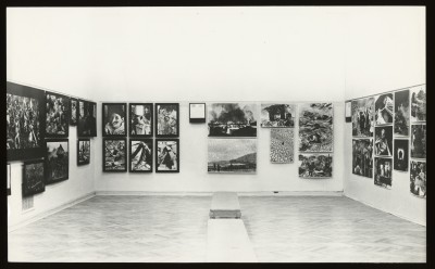 Czarno-białe zdjęcie. W sali wystawy dwóch mężczyzn stoi obok siebie. W tle trzeci, w dłoni trzyma reklamówkę firmy Agfa. W tle obrazy.
