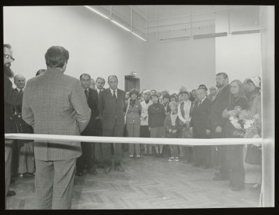 Czarno-białe zdjęcie. Na pierwszym planie biało-czerwona wstęga. Przy niej mężczyzna stojący tyłem, w tle tłum ludzi w sali wystawowej.