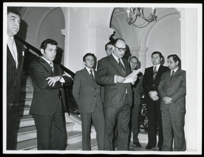 Czarno-białe zdjęcie. Grupka mężczyzn w garniturach na schodach. Mężczyzna pośrodku w okularach, czyta z kartki.