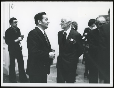 Czarno-białe zdjęcie. Kilka osób w przestrzeni wystawy. W centrum trzy osoby patrzą w różnych kierunkach. W tle zwiedzający i obrazy na ścianie.