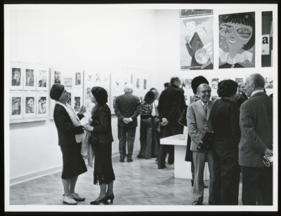 Czarno-białe zdjęcie. W przestrzeni wystawy trzy kobiety i mężczyzna rozmawiają w grupie. Po lewej kobieta tyłem, przygląda się obrazom.