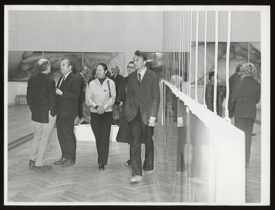Czarno-białe zdjęcie. Na schodach kilka osób. W środku mężczyzna i kobieta ubrana na biało. Z lewej strony ktoś trzyma mikrofon na statywie.