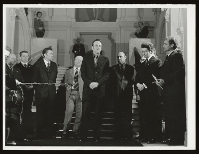 Czarno-białe zdjęcie. Kilkunastu mężczyzn na schodach we wnętrzu Zachęty. Mężczyzna na środku przemawia, inny nagrywa go mikrofonem.
