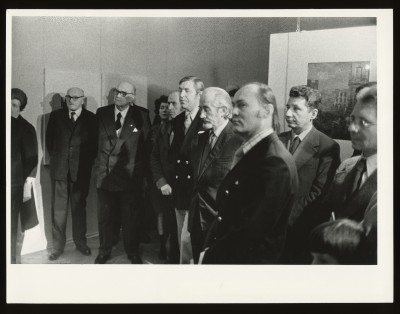 Czarno-białe zdjęcie. Duża grupa ludzi w przestrzeni wystawy. Na środku dwóch mężczyzn w garniturach ściska sobie dłonie.